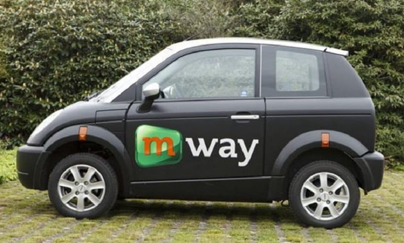 Kleines Elektroauto mit "m-way" Logo, geparkt auf grünem Untergrund, umgeben von Bäumen.